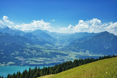 Açık mavi gökyüzünün altında turkuaz bir göl bulunan bir dağ sırasının panoramik görüntüsü.