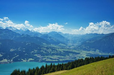 Açık mavi gökyüzünün altında turkuaz bir göl bulunan bir dağ sırasının panoramik görüntüsü.