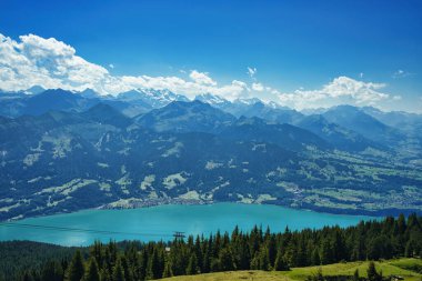 Açık mavi gökyüzünün altında turkuaz bir göl bulunan bir dağ sırasının panoramik görüntüsü.
