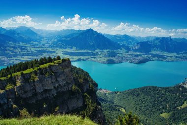 Açık mavi gökyüzünün altında turkuaz bir göl bulunan bir dağ sırasının panoramik görüntüsü.