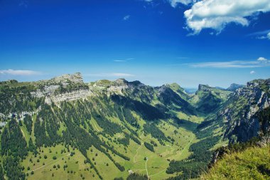 Vadisi olan bir dağ manzarası, yeşil tepeler ve arka planda mavi bir gökyüzü.