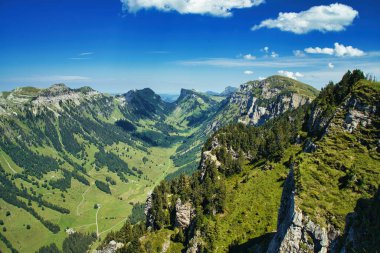Görkemli bir dağ silsilesi, bereketli yeşilliklerle kaplı, açık mavi gökyüzü altında, dağınık bulutlarla, doğanın güzelliğini gözler önüne seriyor.