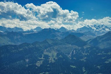 Görkemli bir dağ silsilesi, bereketli yeşilliklerle kaplı, açık mavi gökyüzü altında, dağınık bulutlarla, doğanın güzelliğini gözler önüne seriyor.