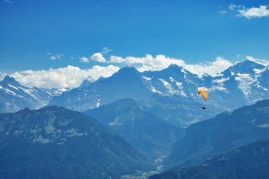 Sersemletici bir dağ manzarasının üzerinde süzülen bir paraglider, açık mavi gökyüzünün altında karla kaplı tepeler ve derin vadilerle
