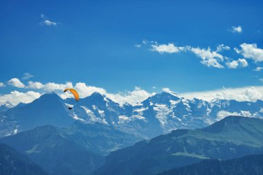 Sersemletici bir dağ manzarasının üzerinde süzülen bir paraglider, açık mavi gökyüzünün altında karla kaplı tepeler ve derin vadilerle