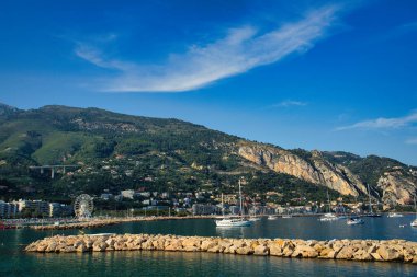 Sahil boyunca renkli binaları olan, berrak bir gökyüzünün altında dağlar tarafından desteklenen sakin bir marina, manzaralı bir sahil manzarası yaratır.