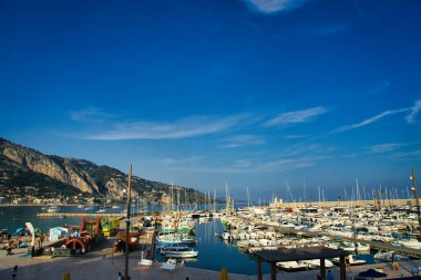 Sahil boyunca renkli binaları olan, berrak bir gökyüzünün altında dağlar tarafından desteklenen sakin bir marina, manzaralı bir sahil manzarası yaratır.