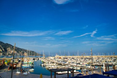 Sahil boyunca renkli binaları olan, berrak bir gökyüzünün altında dağlar tarafından desteklenen sakin bir marina, manzaralı bir sahil manzarası yaratır.