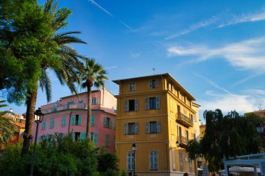 Yat ve teknelerle dolu hareketli bir liman, parçalı bulutlu bir gökyüzünün altındaki renkli tepe binalarının zeminine kurulmuş.
