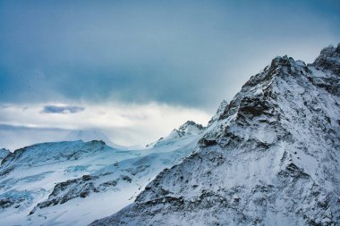 Engebeli zirveleri ve dramatik bulutlu gökyüzü olan görkemli karla kaplı dağlar.