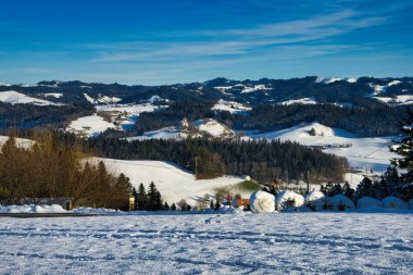 Parlak mavi gökyüzünün altında yeşil ağaçların ve yuvarlanan tepelerin olduğu karlı bir manzara.