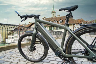 30.05.24. Bern, Swizerland. Üzerinde tarihi bir Avrupa kilisesi ve tarihi bir kasaba mimarisi olan kaldırım taşı köprüsü üzerinde parlak bir elektrikli bisiklet..
