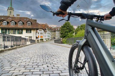 30.05.24. Bern, Swizerland. Üzerinde tarihi bir Avrupa kilisesi ve tarihi bir kasaba mimarisi olan kaldırım taşı köprüsü üzerinde parlak bir elektrikli bisiklet..