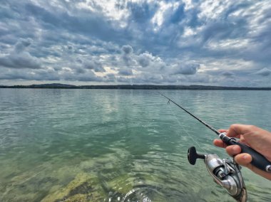 Bulutlu bir gökyüzünün altında sakin turkuaz suların üzerinde bir olta tutan bir elle balık tutma sahnesi huzurlu ve sakin bir atmosfer yaratıyor.