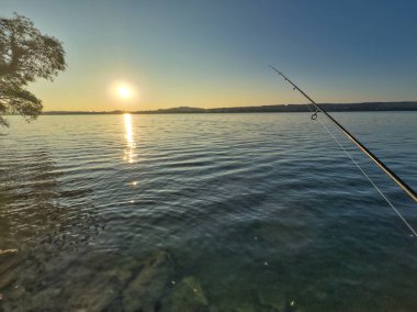 Bulutlu bir gökyüzünün altında sakin turkuaz suların üzerinde bir olta tutan bir elle balık tutma sahnesi huzurlu ve sakin bir atmosfer yaratıyor.