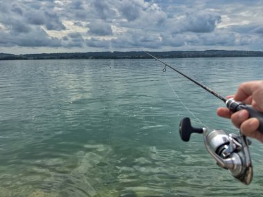 Bulutlu bir gökyüzünün altında sakin turkuaz suların üzerinde bir olta tutan bir elle balık tutma sahnesi huzurlu ve sakin bir atmosfer yaratıyor.