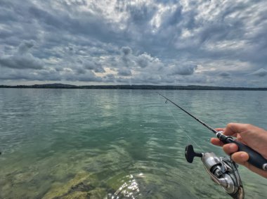 Bulutlu bir gökyüzünün altında sakin turkuaz suların üzerinde bir olta tutan bir elle balık tutma sahnesi huzurlu ve sakin bir atmosfer yaratıyor.