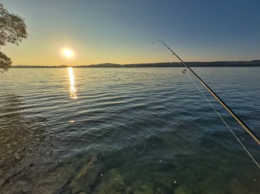 Bulutlu bir gökyüzünün altında sakin turkuaz suların üzerinde bir olta tutan bir elle balık tutma sahnesi huzurlu ve sakin bir atmosfer yaratıyor.