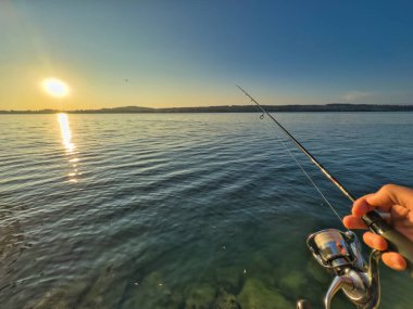 Bulutlu bir gökyüzünün altında sakin turkuaz suların üzerinde bir olta tutan bir elle balık tutma sahnesi huzurlu ve sakin bir atmosfer yaratıyor.