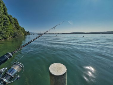 Bulutlu bir gökyüzünün altında sakin turkuaz suların üzerinde bir olta tutan bir elle balık tutma sahnesi huzurlu ve sakin bir atmosfer yaratıyor.