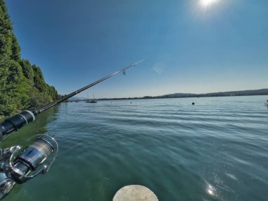 Bulutlu bir gökyüzünün altında sakin turkuaz suların üzerinde bir olta tutan bir elle balık tutma sahnesi huzurlu ve sakin bir atmosfer yaratıyor.