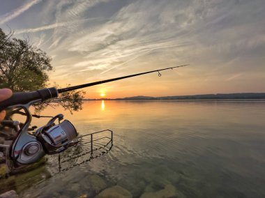 Bulutlu bir gökyüzünün altında sakin turkuaz suların üzerinde bir olta tutan bir elle balık tutma sahnesi huzurlu ve sakin bir atmosfer yaratıyor.