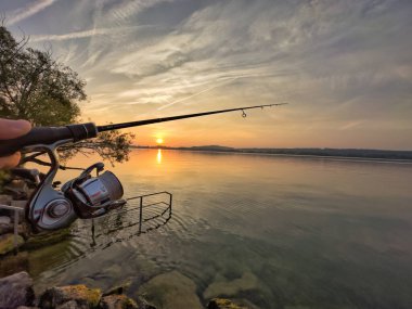 Bulutlu bir gökyüzünün altında sakin turkuaz suların üzerinde bir olta tutan bir elle balık tutma sahnesi huzurlu ve sakin bir atmosfer yaratıyor.