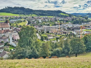 Yuvarlanan tepeler, yeşil otlaklar ve dağılmış ağaçlarla çevrili küçük bir kasabanın manzarası dramatik bulutlu bir gökyüzünün altında güneş parlıyor.