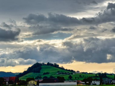 Ağaçlarla ve evlerle kaplı, bulutlu bir gökyüzünün altında, yumuşak akşam ışığıyla aydınlatılmış bir tepe manzaralı dramatik bir kır manzarası..