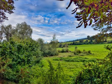 Ağaçlarla ve evlerle kaplı, bulutlu bir gökyüzünün altında, yumuşak akşam ışığıyla aydınlatılmış bir tepe manzaralı dramatik bir kır manzarası..