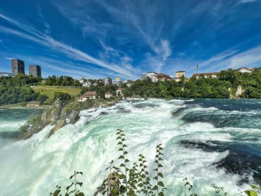Güçlü bir şelale, uçurumlar ve yeşil gökyüzü altında yükselen sis ile dinamik ve dramatik bir manzara yaratıyor. Rhine Falls.