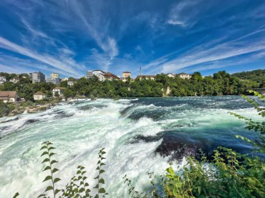 Güçlü bir şelale, uçurumlar ve yeşil gökyüzü altında yükselen sis ile dinamik ve dramatik bir manzara yaratıyor. Rhine Falls.