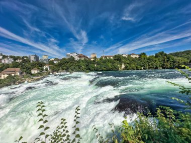 Güçlü bir şelale, uçurumlar ve yeşil gökyüzü altında yükselen sis ile dinamik ve dramatik bir manzara yaratıyor. Rhine Falls.