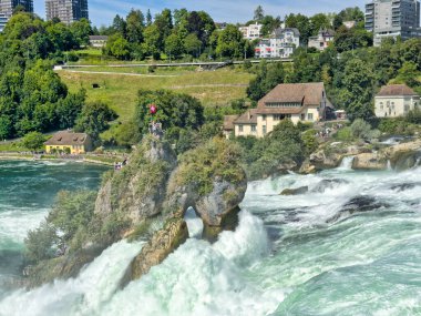 Güçlü bir şelale, uçurumlar ve yeşil gökyüzü altında yükselen sis ile dinamik ve dramatik bir manzara yaratıyor. Rhine Falls.