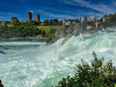 Güçlü bir şelale, uçurumlar ve yeşil gökyüzü altında yükselen sis ile dinamik ve dramatik bir manzara yaratıyor. Rhine Falls.