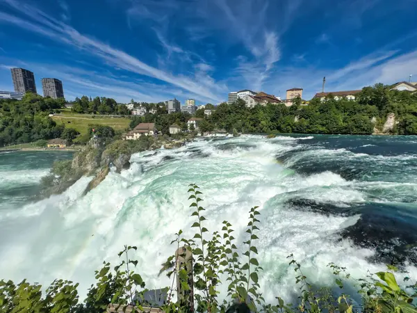 Güçlü bir şelale, uçurumlar ve yeşil gökyüzü altında yükselen sis ile dinamik ve dramatik bir manzara yaratıyor. Rhine Falls.