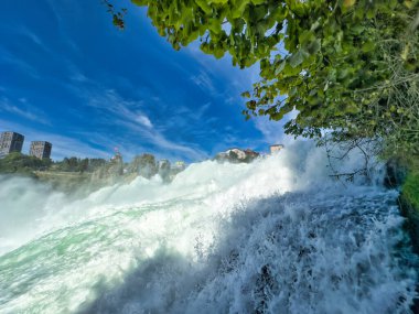 Güçlü bir şelale, uçurumlar ve yeşil gökyüzü altında yükselen sis ile dinamik ve dramatik bir manzara yaratıyor. Rhine Falls.
