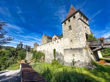 Tarihsel taş kale duvarı ve kulesi parlak mavi gökyüzünün altında yemyeşil bir yeşillikle çevrili, ortaçağ mimarisi ve manzaralı bir güzellik sergiliyor.