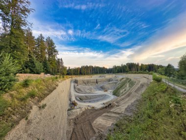 Gece gökyüzünün altında yoğun yeşil ormanlarla çevrili bir taş ocağında inşaat araçları görülebiliyor.