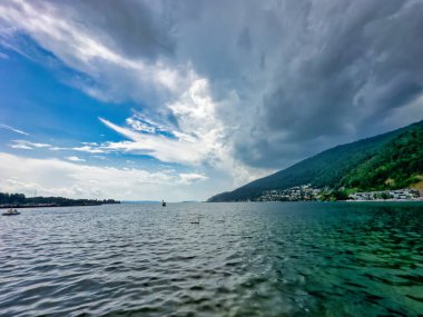 Sakin sularla huzurlu göl manzarası, uzak bir heykel ve parçalı bulutlu bir gökyüzünün altında sakin bir ufuk.