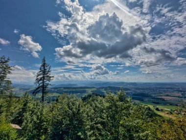 Yeşil yemyeşil bir vadinin panoramik manzarası ve dramatik bir gökyüzünün altındaki kayan tepeler dağınık bulutlarla dolu, doğanın güzelliğini gösteriyor.