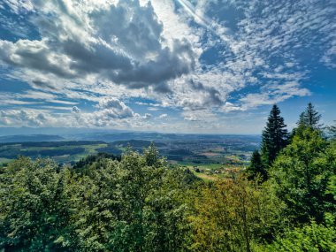 Yeşil yemyeşil bir vadinin panoramik manzarası ve dramatik bir gökyüzünün altındaki kayan tepeler dağınık bulutlarla dolu, doğanın güzelliğini gösteriyor.