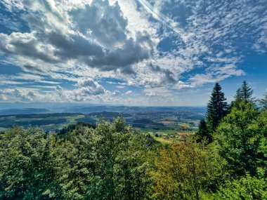 Yeşil yemyeşil bir vadinin panoramik manzarası ve dramatik bir gökyüzünün altındaki kayan tepeler dağınık bulutlarla dolu, doğanın güzelliğini gösteriyor.