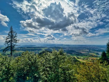 Yeşil yemyeşil bir vadinin panoramik manzarası ve dramatik bir gökyüzünün altındaki kayan tepeler dağınık bulutlarla dolu, doğanın güzelliğini gösteriyor.