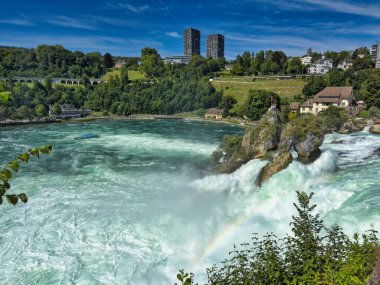 Güçlü bir şelale, uçurumlar ve yeşil gökyüzü altında yükselen sis ile dinamik ve dramatik bir manzara yaratıyor. Rhine Falls.