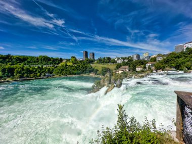 Güçlü bir şelale, uçurumlar ve yeşil gökyüzü altında yükselen sis ile dinamik ve dramatik bir manzara yaratıyor. Rhine Falls.