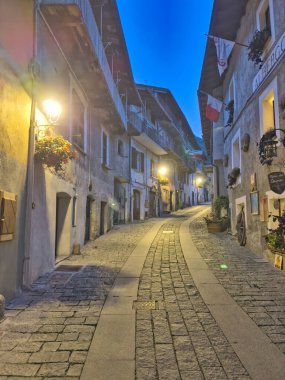 Dağlık bir köyde sakin bir alacakaranlık manzarası. Kırsal evler, parlayan ışıklar ve derin mavi bir gökyüzünün altında yükselen tepeler..