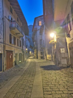 Dağlık bir köyde sakin bir alacakaranlık manzarası. Kırsal evler, parlayan ışıklar ve derin mavi bir gökyüzünün altında yükselen tepeler..