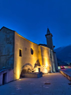 Dağlık bir köyde sakin bir alacakaranlık manzarası. Kırsal evler, parlayan ışıklar ve derin mavi bir gökyüzünün altında yükselen tepeler..