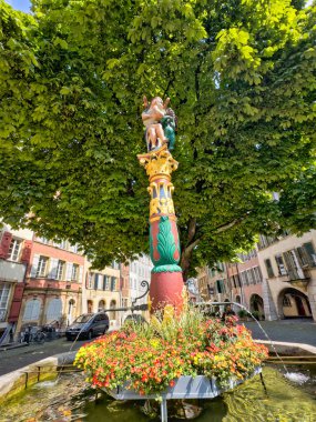 Renkli bir heykel, bir saat kulesi ve parçalı bulutlu mavi gökyüzünün altında karmaşık detaylar içeren ortaçağ mimarisi.